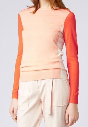 Kobieta nosząca sweter z długim rękawem w stylu color-block z jasnoróżową przednią częścią i jaskrawo pomarańczowymi rękawami, połączony z kremowymi spodniami z wysokim stanem.