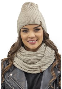 Beige strikket hue med ribbet design og brun lædermærket, parret med et matchende struktureret tørklæde, båret over en sort læderjakke.