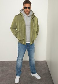 Giacca imbottita verde oliva con cappuccio, felpa grigia sotto, jeans blu strappati e sneakers bianche. Accessoriata con occhiali da sole neri.
