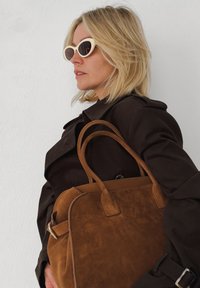 Bruine suède tote bag met leren accenten en twee korte handvatten, gecombineerd met een donkere trenchcoat en lichtgekleurde zonnebril.