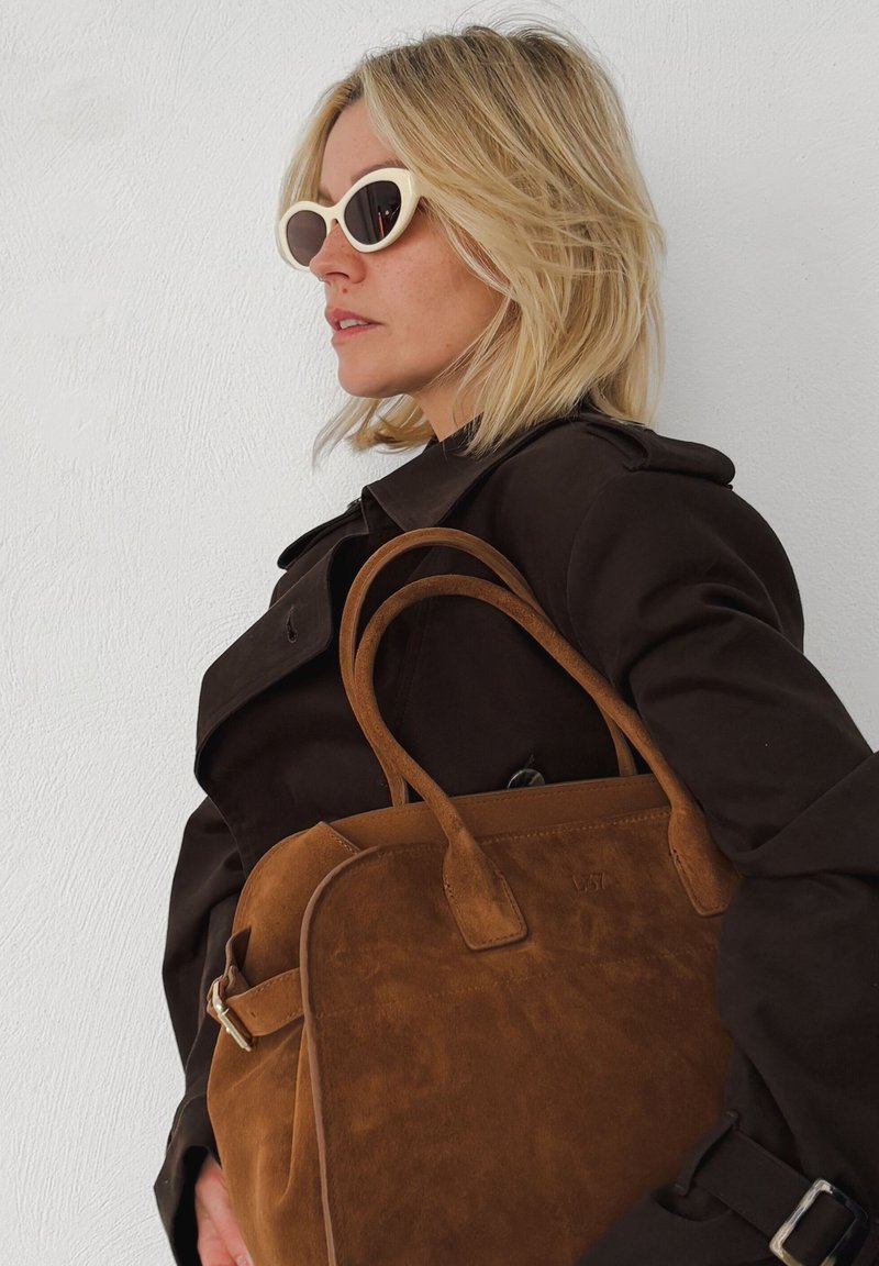 Bruine suède tote bag met leren accenten en twee korte handvatten, gecombineerd met een donkere trenchcoat en lichtgekleurde zonnebril.