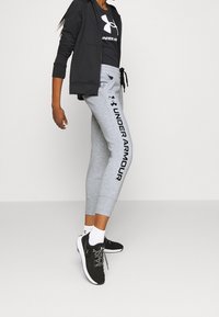 Sudadera con cremallera negra, pantalones de chándal gris ajustados con "UNDER ARMOUR" impreso en negro y zapatillas deportivas negras con cordones grises y suelas blancas.