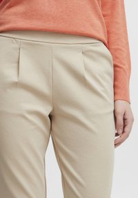 Pantalons plissés beiges en tissu doux, présentant une texture lisse et une ceinture plate. La main du modèle est posée sur la hanche.