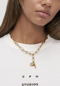 Guldmetal kædethalskæde med store led, en vedhæng med et graveret logo, båret over en hvid T-shirt med sort tekst.