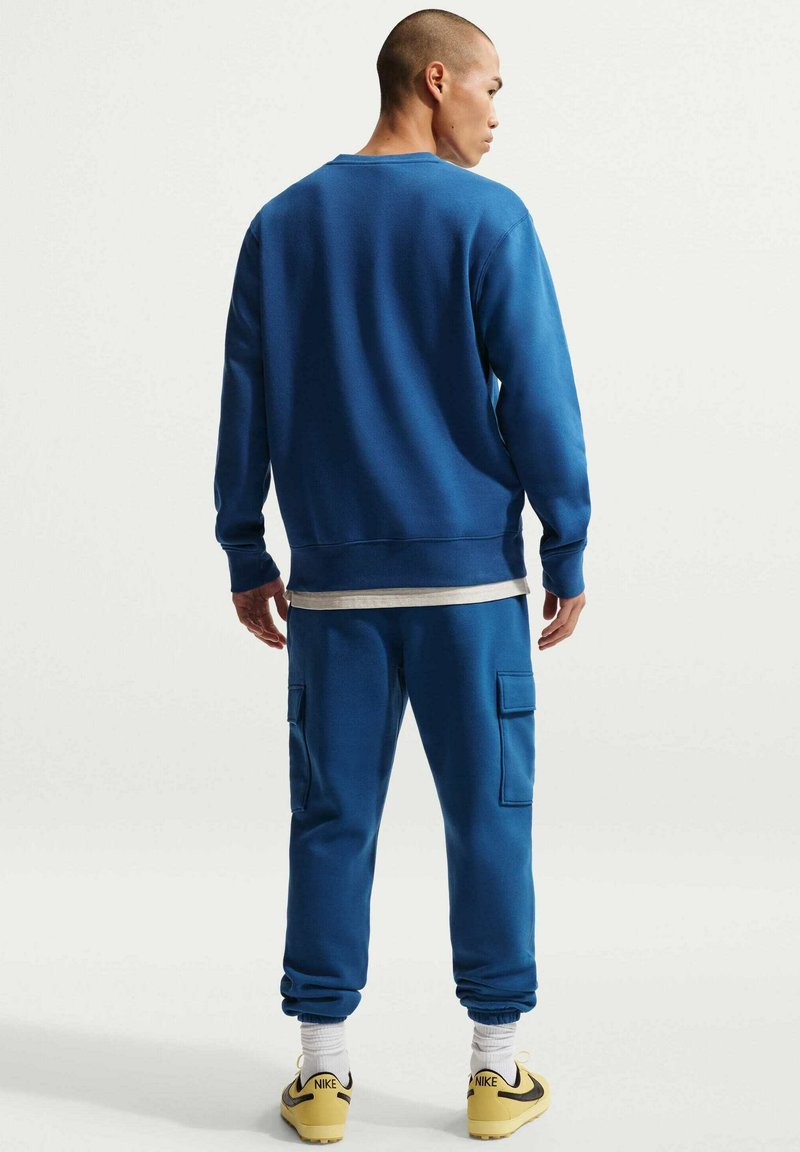 Sweat-shirt bleu à col rond, poignets côtelés, et pantalon de survêtement bleu assorti avec poches cargo. Porté avec des baskets jaunes.