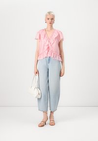 Blusa rosa a volant con maniche corte, abbinata a jeans a vita alta azzurri e sandali con tacco beige. Modella che tiene una borsa bianca.