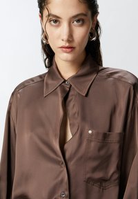 Smeđi svilen košulja s kragnom na dugmad, jednim džepom na prsima i suptilnim sjajem. Ima opuštenu kroj i pola otvoren dizajn.