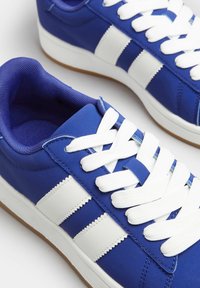 Niebieskie trampki z białymi paskami i sznurówkami. Gładka syntetyczna cholewka i gumowa podeszwa. Posiadają wyściełany kołnierz i teksturowane akcenty.