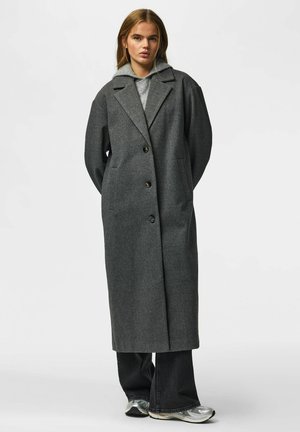 Jeune femme portant un long manteau gris foncé sur un sweat à capuche gris, un pantalon large noir et des baskets argentées, debout devant un fond uni.