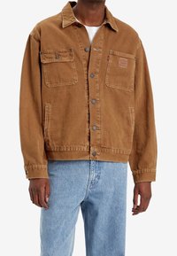 Mężczyzna w brązowej kurtce zapinanej na guziki z dwoma kieszeniami na piersi oraz jasnoniebieskich dżinsach, stojący na białym tle.
