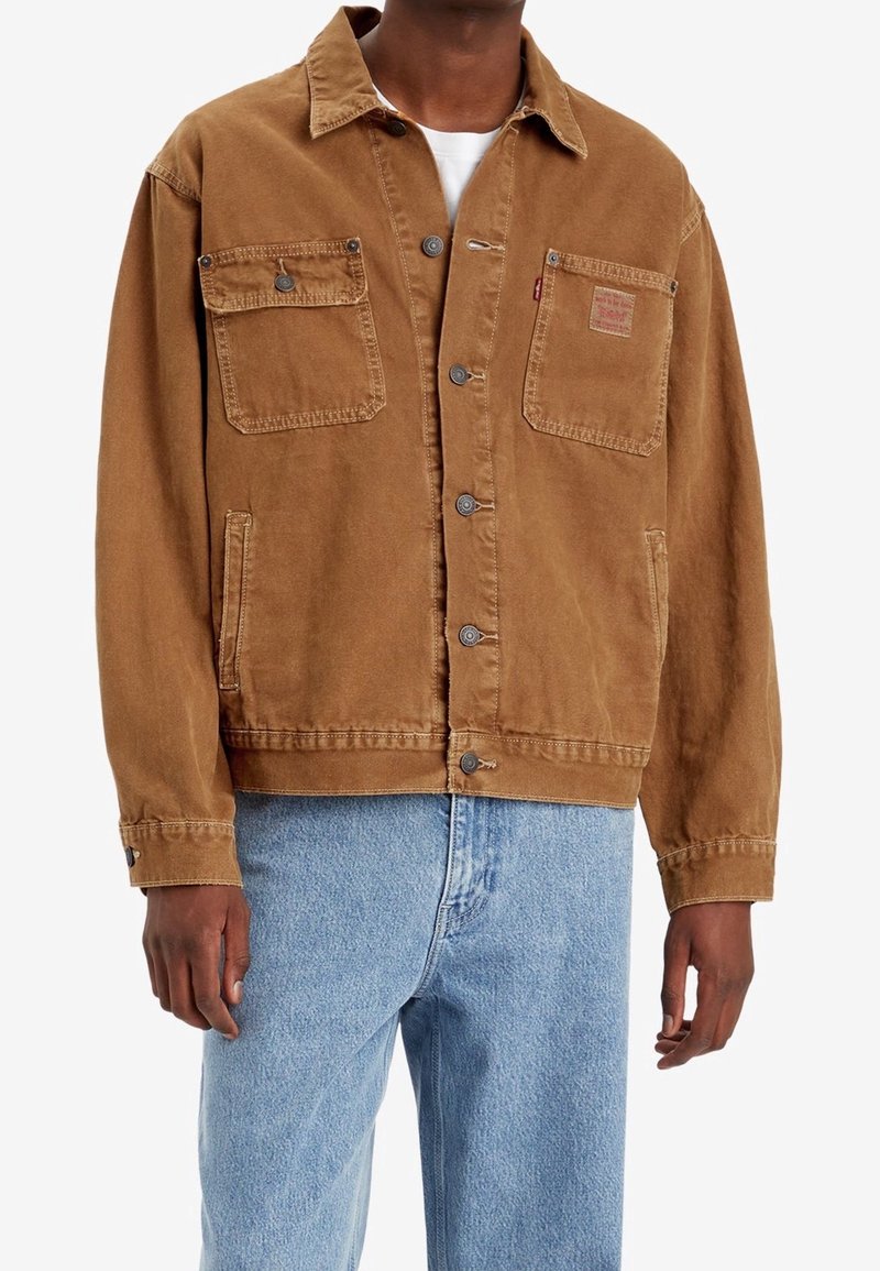 Mężczyzna w brązowej kurtce zapinanej na guziki z dwoma kieszeniami na piersi oraz jasnoniebieskich dżinsach, stojący na białym tle.