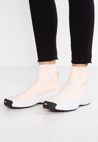 Lichtroze sokachtige sneakers hebben een rekbare textiele bovenkant, een zwarte zool en een logoaccent op de hiel en de teen. Textuur van de zwarte buitenzool.