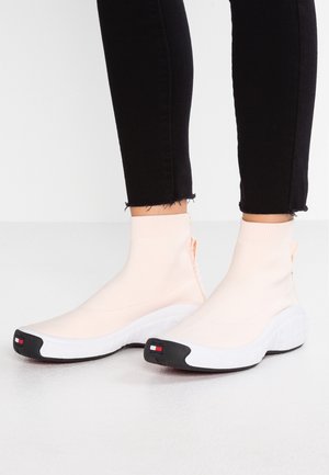 Lichtroze sokachtige sneakers hebben een rekbare textiele bovenkant, een zwarte zool en een logoaccent op de hiel en de teen. Textuur van de zwarte buitenzool.