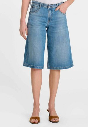 Persona che indossa pantaloncini di jeans azzurri fino al ginocchio con mano in tasca e sandali marroni con punta aperta su sfondo bianco.