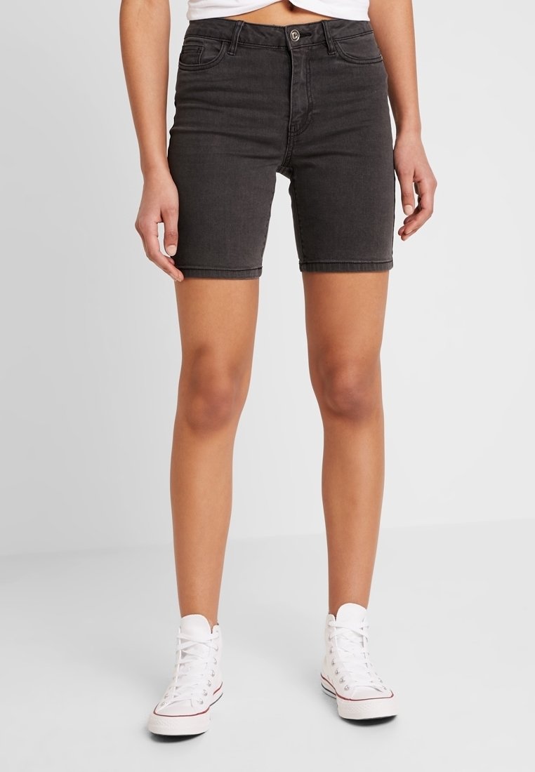 Personne portant un short en jean noir taille haute et des baskets montantes blanches, debout devant un fond uni.