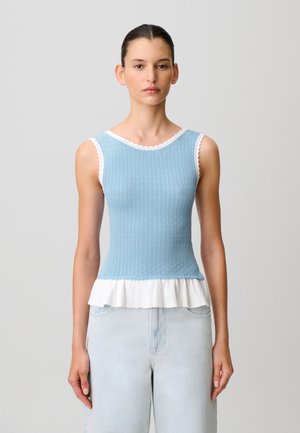Donna che indossa un top senza maniche in maglia azzurro chiaro con bordi bianchi e orlo arricciato, abbinato a jeans azzurri, in piedi davanti a uno sfondo semplice.