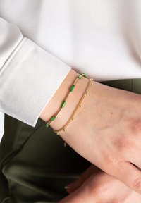 Zestaw bransoletek w kolorze złotym i zielonym, składający się z cienkich łańcuszków i małych dekoracyjnych koralików, noszony na nadgarstku z lekką tkaninową rękawiczką w tle.