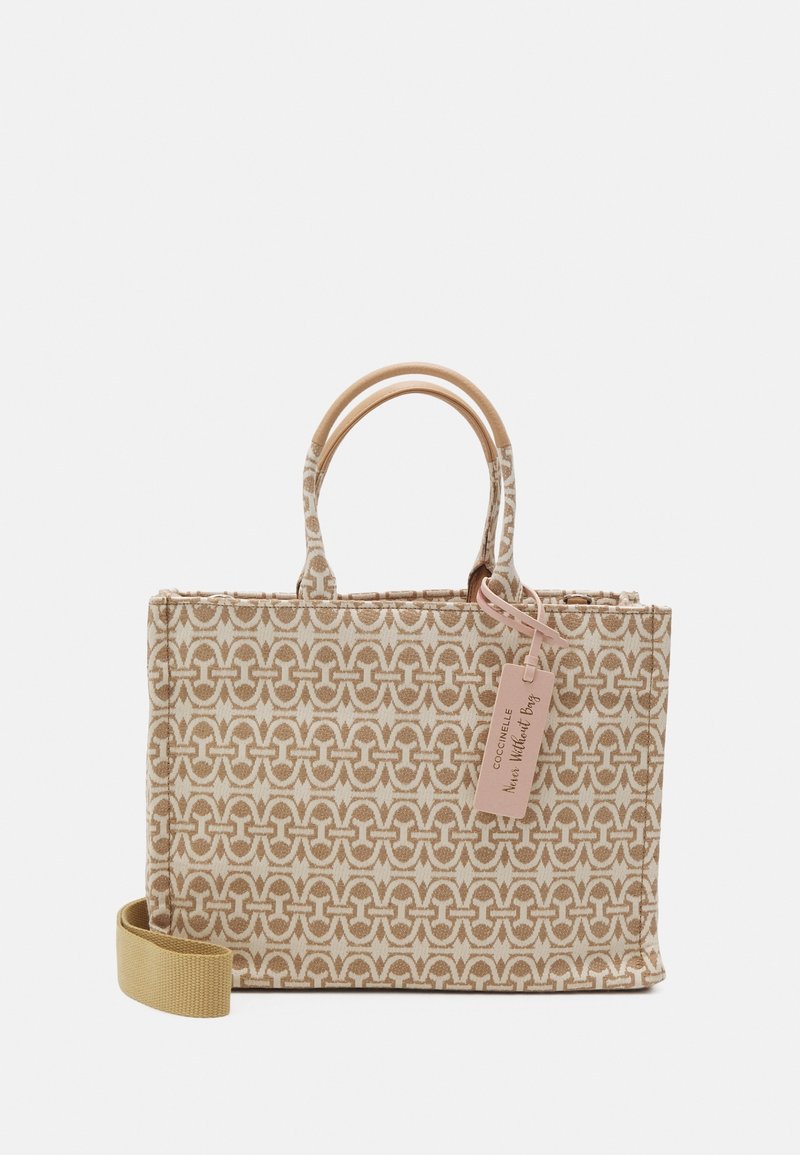 Beżowa torba materiałowa z geometrycznym wzorem, z podwójnymi rączkami u góry i odpinanym paskiem. Zawiera metkę z informacjami o marce.