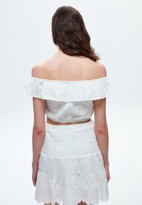 Femme aux cheveux bruns ondulés portant un haut blanc en dentelle ajourée décolleté épaule et une jupe assortie, debout de dos face à l'appareil photo.