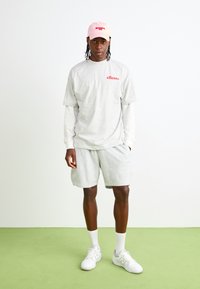 Sivý tričko s červeným logom "ellesse", spárované so sivými šortkami. Rúžová čiapka, biele ponožky a biele tenisky. Jednoduchý, športový outfit na zelenej podlahe.