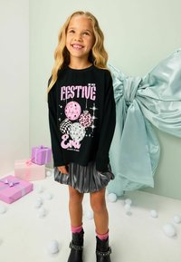 Čierna košeľa s dlhým rukávom a farebnou grafikou balónov, text "Festive Era", v kombinácii so šedou lesklou sukňou, ružovými ponožkami a čiernymi čižmami.