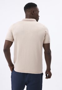 Camisa polo de manga corta beige con una textura sutil y un cuello clásico. Presenta detalles de rayas en contraste cerca del cuello y un ajuste relajado.