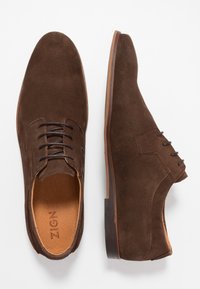 Scarpe in suede marrone con una texture liscia, dotate di punta arrotondata, sistema di allacciatura e suola in un marrone chiaro contrastante.