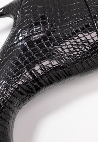 Material patent negro con relieve de cocodrilo y textura brillante, moldeado en un diseño contorneado con un borde curvado y detalles de costura.