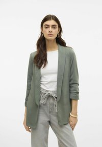 Groene zachte stoffen blazer met een open voorkant, lange mouwen en een losse pasvorm. Gekleed over een witte top en gestyled met lichtgrijze broeken.