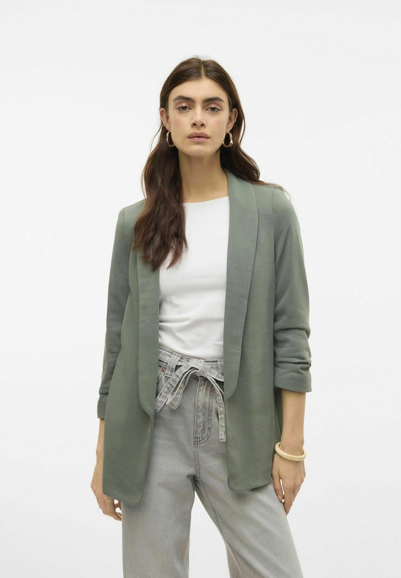 Groene zachte stoffen blazer met een open voorkant, lange mouwen en een losse pasvorm. Gekleed over een witte top en gestyled met lichtgrijze broeken.
