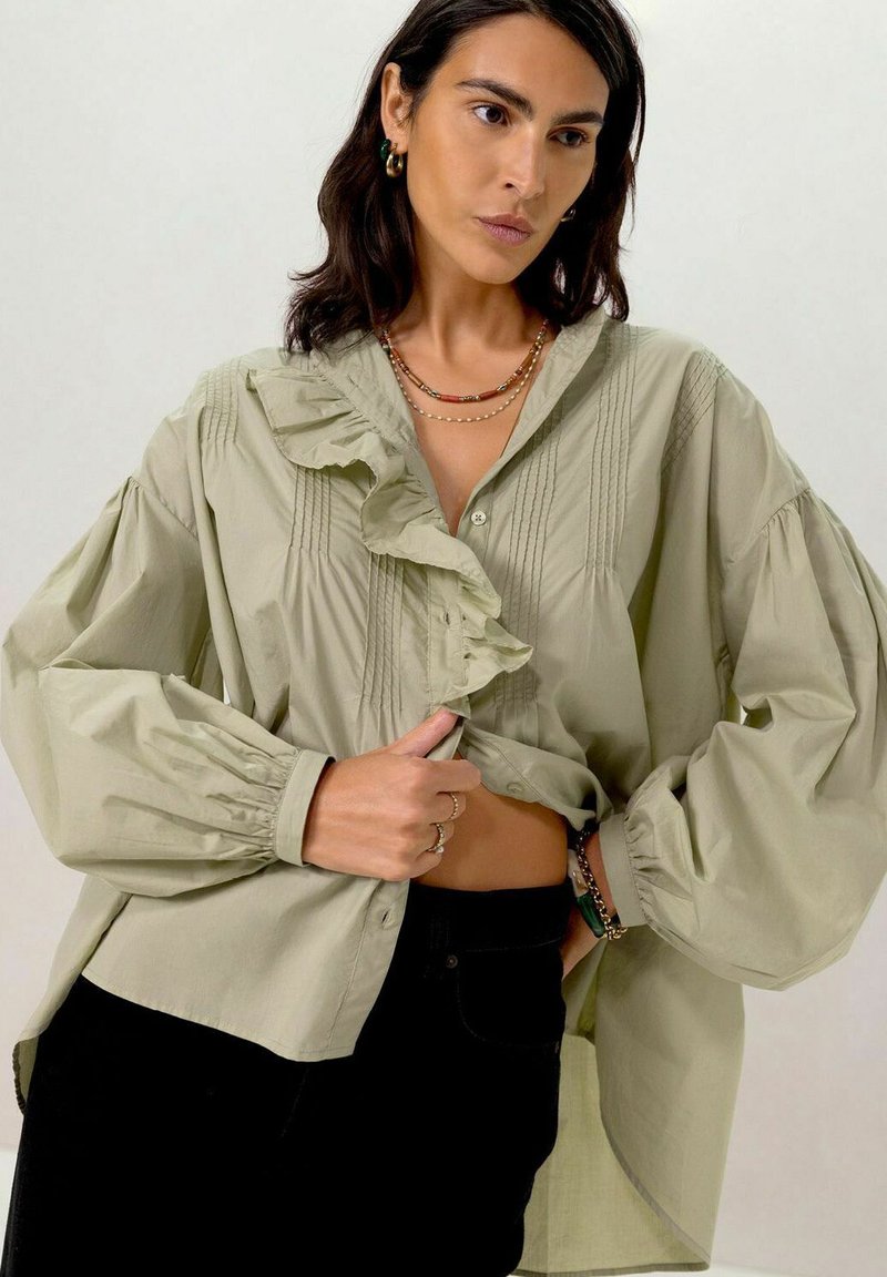 Chemise boutonnée vert olive clair avec des détails froncés, des plis et des manches bouffantes surdimensionnées. Associée à un jean noir.
