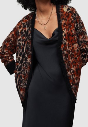 Femme portant un cardigan duveteux à motif léopard rouge et noir sur une robe noire en satin, colliers en or superposés, aucun arrière-plan visible.