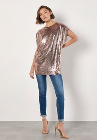 Top de lentejuelas en rosa dorado brillante con mangas cortas y anchas; combinado con jeans azules ajustados y tacones nude con tiras. Textura lisa y brillante.
