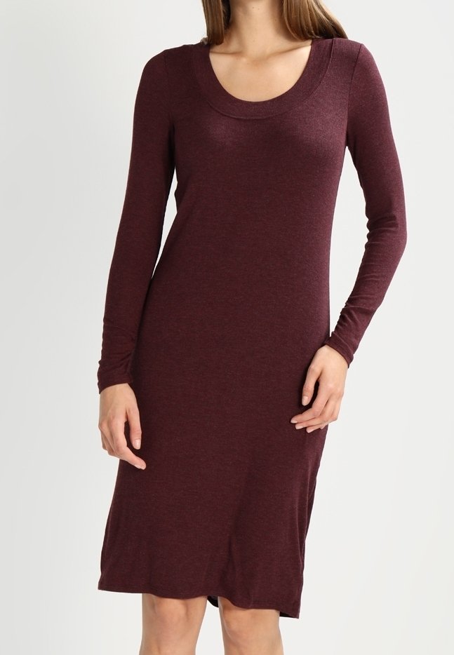 Femme portant une robe ajustée bordeaux à manches longues et longueur genou, avec un col rond, debout devant un fond clair uni.