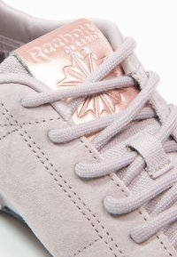 Gros plan d'une basket Reebok Classic en daim violet clair avec des lacets assortis et un logo métallique en or rose sur la languette.