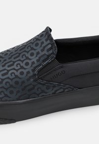 Musta liukusovittava kengät tekstuurilla varustetusta päällisestä, jossa on sävyyn sointuva kuviolaatta. Varustettu joustavilla sivupaneeleilla ja yhtenäisellä mustalla kumipohjalla.