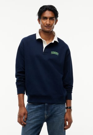 Man draagt een marineblauwe polotrui met lange mouwen, witte kraag en tekst "TOMMY", gecombineerd met een blauwe spijkerbroek en een zwart polshorloge.