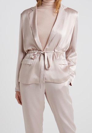 Kobieta ubrana w jasnoróżową satynową kurtkę z paskiem do wiązania, do kompletu spodnie oraz jasnoróżowy prążkowany sweter z golfem, ręce swobodnie opadające wzdłuż ciała.
