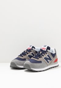 Un paio di scarpe New Balance 574 grigio e blu navy con dettagli rossi, in mesh e suede, posizionate su una superficie bianca.