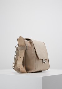 Béžová plátěná taška přes rameno s klopou, texturovaným povrchem a řetízkovým popruhem; obsahuje kovové akcenty.