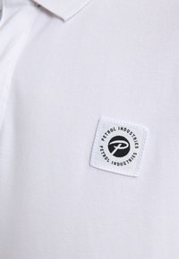 Polo shirt blanc en tissu texturé, avec une étiquette tissée sur la poitrine affichant "Petrol Industries" en noir et blanc.