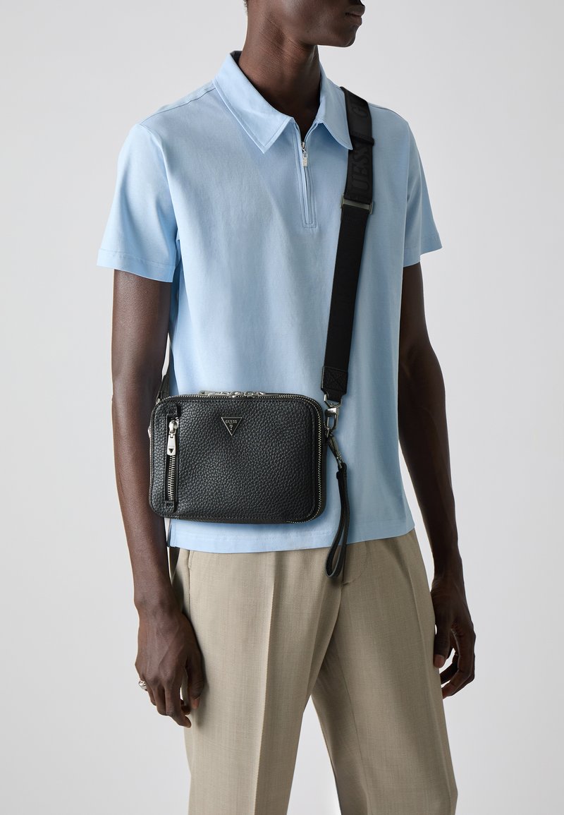 Bolso cruzado de cuero negro texturizado con cierre de cremallera, herrajes plateados y correa ajustable, combinado con una camiseta polo azul claro y pantalones beiges.