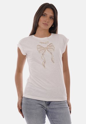 Giovane donna che indossa una t-shirt bianca con maniche arrotolate, decorata con un disegno di fiocco dorato e la scritta "YES ZEE", abbinata a jeans azzurri.