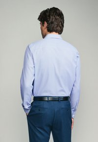 Chemise bleu clair à boutons avec un motif à petits points subtil, associée à un pantalon bleu marine et une ceinture noire, vue de dos.