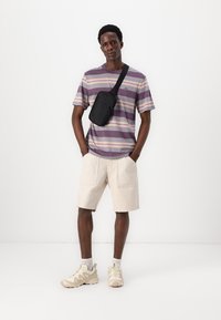 Pruhovaná košile s krátkým rukávem v purpurové a béžové barvě, béžové šortky a neutrální tenisky. Černá crossbody taška nošená na rameni.