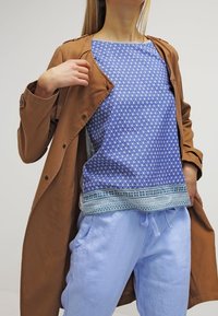 Abrigo marrón abotonado sobre una blusa azul con estampado de formas geométricas y un dobladillo decorativo, combinado con pantalones azul claro.