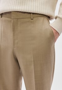 Μπεζ παντελόνια ραμμένα με προσοχή, με �λεία υφή, πλευρικές τσέπες και κομψή εφαρμογή. Συνδυάζονται με ένα ελαφρύ πουλόβερ με ραβδώσεις πάνω από αυτά.