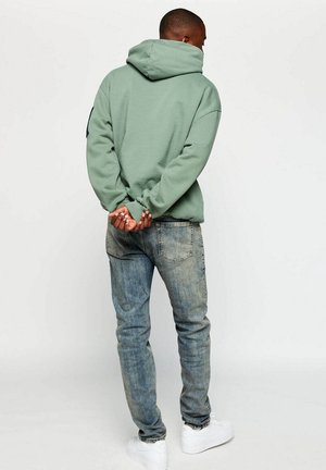 Mand i lysegrøn hættetrøje, blå jeans og hvide sneakers stående med hænderne foldet bag ryggen mod en ensfarvet baggrund.