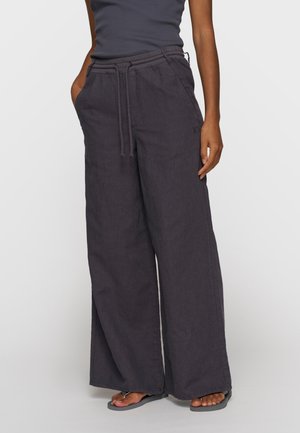 Donna che indossa pantaloni larghi e morbidi di colore grigio scuro con vita con coulisse, top scuro senza maniche e sandali, in piedi con le mani nelle tasche.