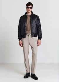 Schwarze Bomberjacke mit strukturierter Oberfläche, beiger gestrickter Pullover, beige Hose und schwarze Lederstiefel. Accessoires umfassen schwarze Sonnenbrillen.